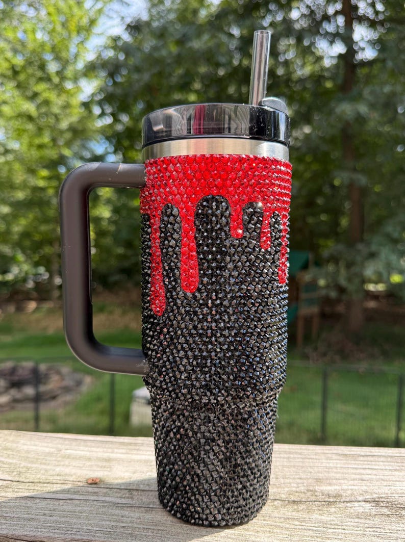 Puede incluir: Un vaso adornado con pedrer&iacute;a con asa y tapa negras. La secci&oacute;n superior est&aacute; cubierta de pedrer&iacute;a roja, con un dise&ntilde;o de goteo de pedrer&iacute;a negra que cae en cascada por el cuerpo. Una pajita plateada est&aacute; insertada en la tapa.