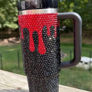 Puede incluir: Taza de viaje adornada con pedrer&iacute;a negra y roja. Las piedras rojas est&aacute;n dispuestas para que parezcan gotas sobre las negras. La taza tiene un asa y una tapa negras, y una pajita plateada.