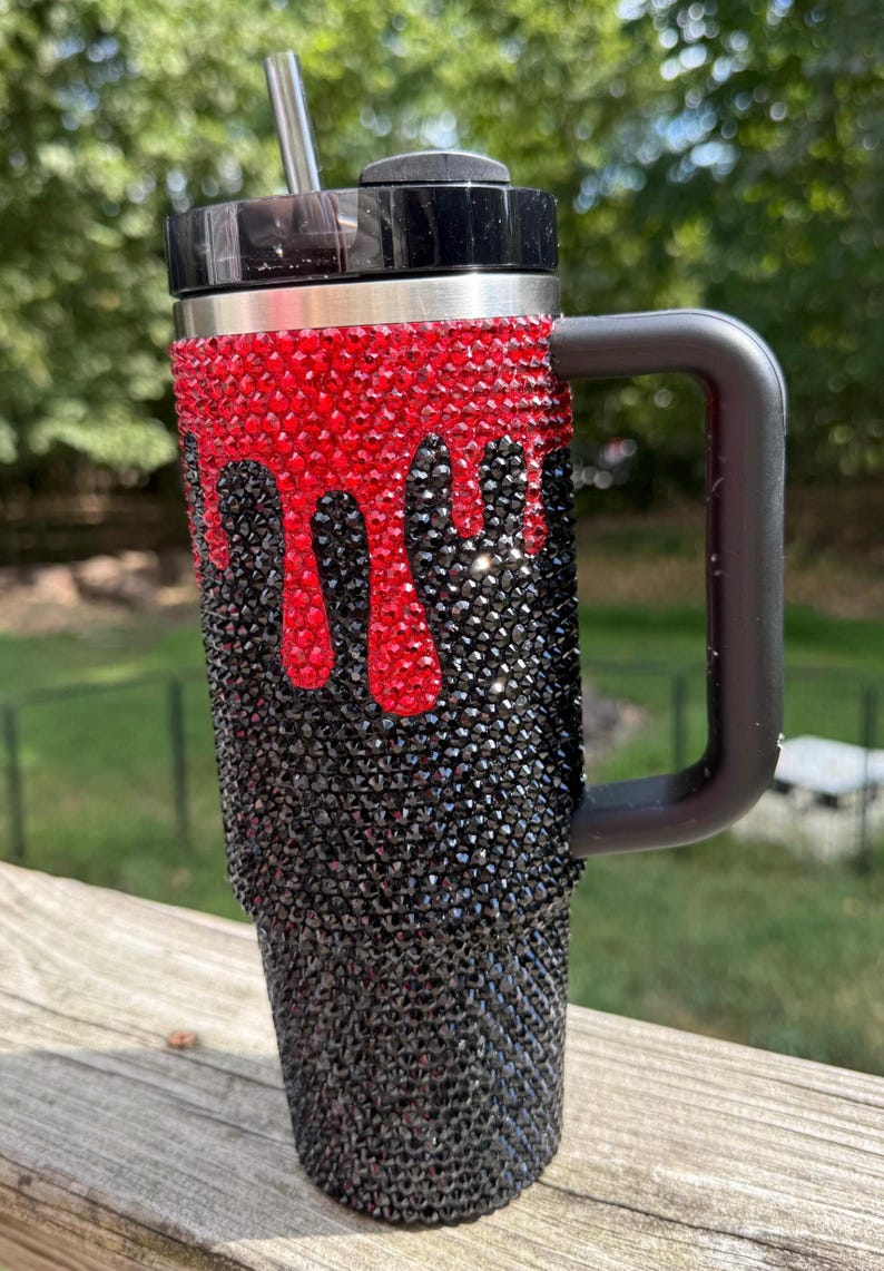 Puede incluir: Taza de viaje adornada con pedrer&iacute;a negra y roja. Las piedras rojas est&aacute;n dispuestas para que parezcan gotas sobre las negras. La taza tiene un asa y una tapa negras, y una pajita plateada.