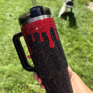Puede incluir: Un vaso negro con asa, adornado con pedrer&iacute;a roja y negra. La pedrer&iacute;a roja est&aacute; dispuesta en un patr&oacute;n de goteo sobre la pedrer&iacute;a negra. El vaso tiene una tapa negra y una pajita transparente.