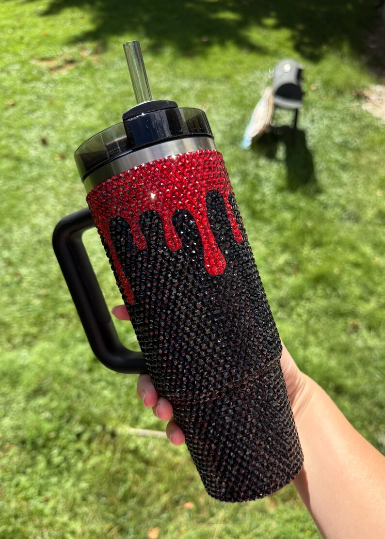Puede incluir: Un vaso negro con asa, adornado con pedrer&iacute;a roja y negra. La pedrer&iacute;a roja est&aacute; dispuesta en un patr&oacute;n de goteo sobre la pedrer&iacute;a negra. El vaso tiene una tapa negra y una pajita transparente.