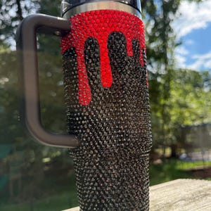Puede incluir: Un vaso negro con asa, cubierto de peque&ntilde;os y brillantes pedrer&iacute;a negra. Pedrer&iacute;a roja gotea desde la parte superior, creando un dise&ntilde;o &uacute;nico. El vaso tiene una tapa negra y una pajita plateada.