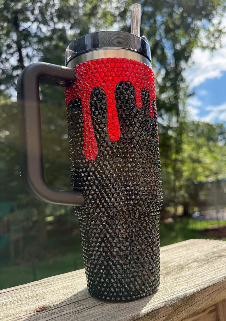 Puede incluir: Un vaso negro con asa, cubierto de peque&ntilde;os y brillantes pedrer&iacute;a negra. Pedrer&iacute;a roja gotea desde la parte superior, creando un dise&ntilde;o &uacute;nico. El vaso tiene una tapa negra y una pajita plateada.