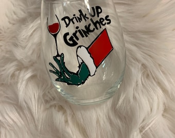 May include: A clear glass wine glass with a green Grinch hand holding a glass of red wine. The glass has the text "Drink Up Grinches" printed on it.