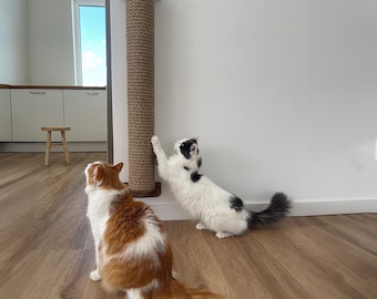 May include: A tall, brown rope cat scratching post mounted on a white wall. Two cats, one orange and white, the other black and white, interact with the post. The post has a dark brown wooden top and bottom.