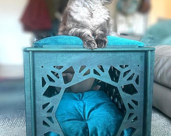 May include: A dark teal wooden pet house with a geometric cutout pattern on the side panels. The structure features a plush, tufted teal velvet cushion inside and a matching teal velvet top surface, with a grey cat resting on the top.