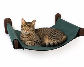 May include: A tabby cat rests on a wall-mounted cat hammock. The hammock is made of a dark green fabric and is supported by wooden brackets. The cat has brown and black stripes and is looking directly at the camera.