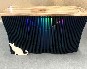 May include: A dark wood table with a unique, wavy design. The table has a light wood top and a black base with a rainbow-colored light shining through the slats. A white wooden cat cutout is placed on the table.