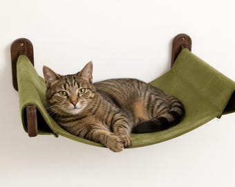 May include: A wall-mounted cat hammock featuring a sage green fabric sling suspended between two dark brown wooden brackets. A brown tabby cat is resting on the fabric surface, which is secured to the wall with visible metal hardware.