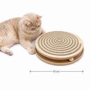 Puede incluir: Un rascador circular de madera para gatos con una superficie de cuerda beige en espiral. La base es de madera clara e incluye una pequeña bola de madera. Un indicador de medición muestra un diámetro de 43 cm.