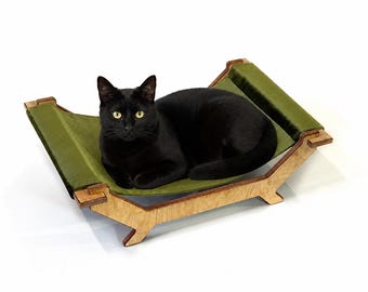 May include: A black cat resting on a pet hammock featuring a light-colored wooden frame with a curved base and a soft olive green fabric bed suspended between two elevated ends. The structure is designed for small animals to lounge comfortably.