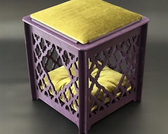 May include: A purple wooden pet bed with a geometric cut-out design. It features a mustard-yellow cushion on top and a matching cushion inside. The bed is cube-shaped and has a modern, decorative style.