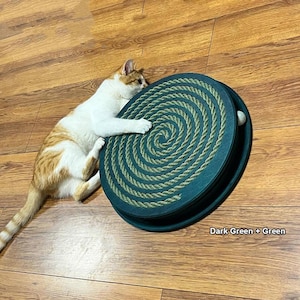 Op de afbeelding: Een donkergroen kattenspeeltje met een spiraalvormig touwontwerp en een witte bal, met een wit en oranje kat ernaast. De tekst "Dark Green + Green" is zichtbaar.
