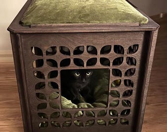 May include: A dark brown wooden cat house with a decorative pattern and an olive green cushion. A black cat is inside the house. The text on the image says "Wood: Belgian Chocolate, Cushions: Olive, Pattern: Sweet Rocket."