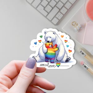 Könnte beinhalten: Ein Aufkleber mit einem weißen Eisbären, der einen Regenbären umarmt. Der Aufkleber hat einen weißen Rand, Regenbogenherzen und den Text "LOVE is love" mit einem Herzsymbol. Der Aufkleber wird von einer Hand gehalten.