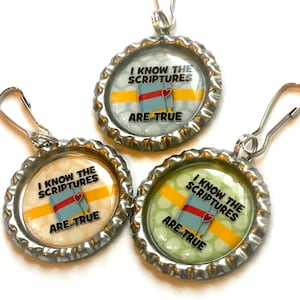 May include: Three silver bottle cap charms with a yellow stripe and white polka dots. Each charm has a blue book with a red heart and the text "I know the scriptures are true".