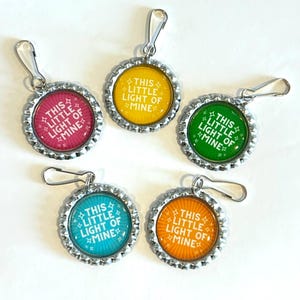 May include: Five bottle cap charms with silver-colored metal edges and attached clasps. Each charm has a different colored background: pink, yellow, green, blue, and orange. The text "THIS LITTLE LIGHT OF MINE" is printed on each charm.