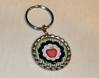 May include: A silver key chain with a bottle cap charm. The charm has a green and black striped background with a black and white scalloped border. A red apple with a green leaf is in the center of the charm.
