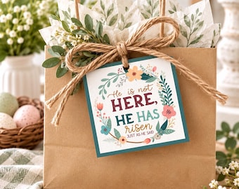 May include: A brown paper gift bag with a square tag featuring a floral wreath and the text He is not HERE, HE HAS RISEN JUST AS HE SAID. The bag is tied with twine and decorated with green foliage and white tissue paper with leaf prints.