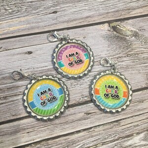 May include: Three silver bottle cap charms with a colorful design and the text "I am a child of God". Each charm has a silver clip attached.