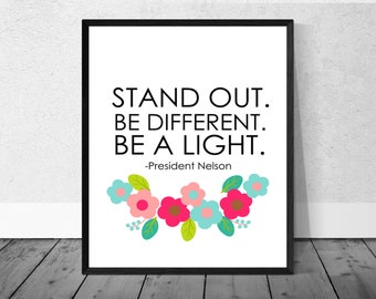以下が含まれることがあります： 白い背景に黒い枠で囲まれたプリント。黒いテキストで「Stand Out. Be Different. Be a Light.」という引用文が書かれている。引用文は「President Nelson」に帰属している。プリントの下部には、カラフルな花が並んでいる。