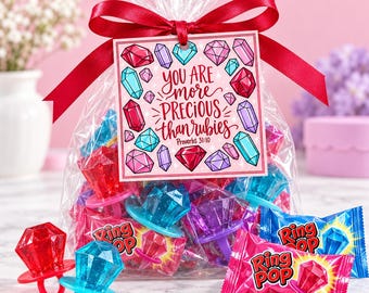 May include: A clear plastic bag filled with assorted colorful Ring Pop candy rings, tied with a red ribbon and a square tag featuring the text You are more precious than rubies Proverbs 31:10 surrounded by illustrated colorful gemstones.