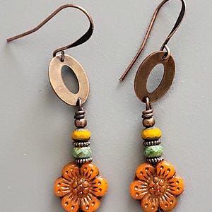 May include: A pair of handcrafted earrings featuring copper-colored metalwork. Each earring has a copper oval, a cluster of yellow and green beads, and an orange flower-shaped bead. The earrings hang from copper ear wires.