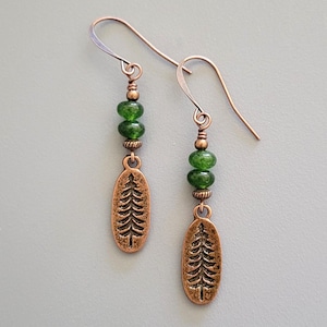 May include: Copper-colored earrings with a tree design. Each earring features a green bead cluster and a copper oval pendant with an embossed tree. The earrings have a hook closure and are displayed against a gray background.