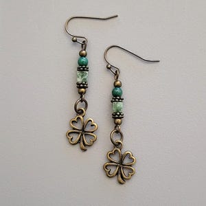 May include: A pair of dangle earrings featuring a four-leaf clover charm. The earrings have a bronze finish with green and speckled beads. The ear wires are bronze-colored.
