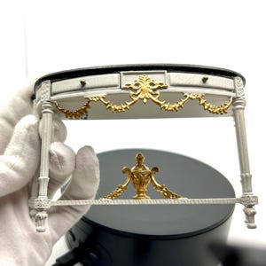 May include: White and gold ornate table with a black top and a decorative urn design on the bottom shelf. The table has a curved top and intricate details.