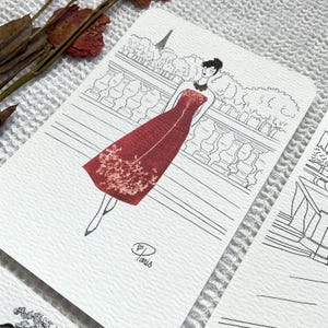 May include: A black and white line drawing of a woman in a red dress standing in front of a Parisian cityscape. The dress is decorated with dried flowers. The text "Paris" is written in cursive at the bottom of the image.