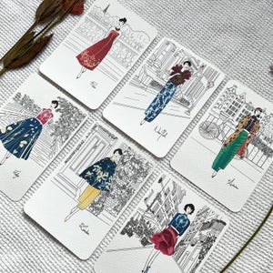 May include: Set of six illustrated cards featuring women in different outfits and locations. Each card has a black and white illustration of a woman in a different city, with the city name written below the illustration. The cities featured are Paris, New York, Amsterdam, London, Tokyo, and Rome.