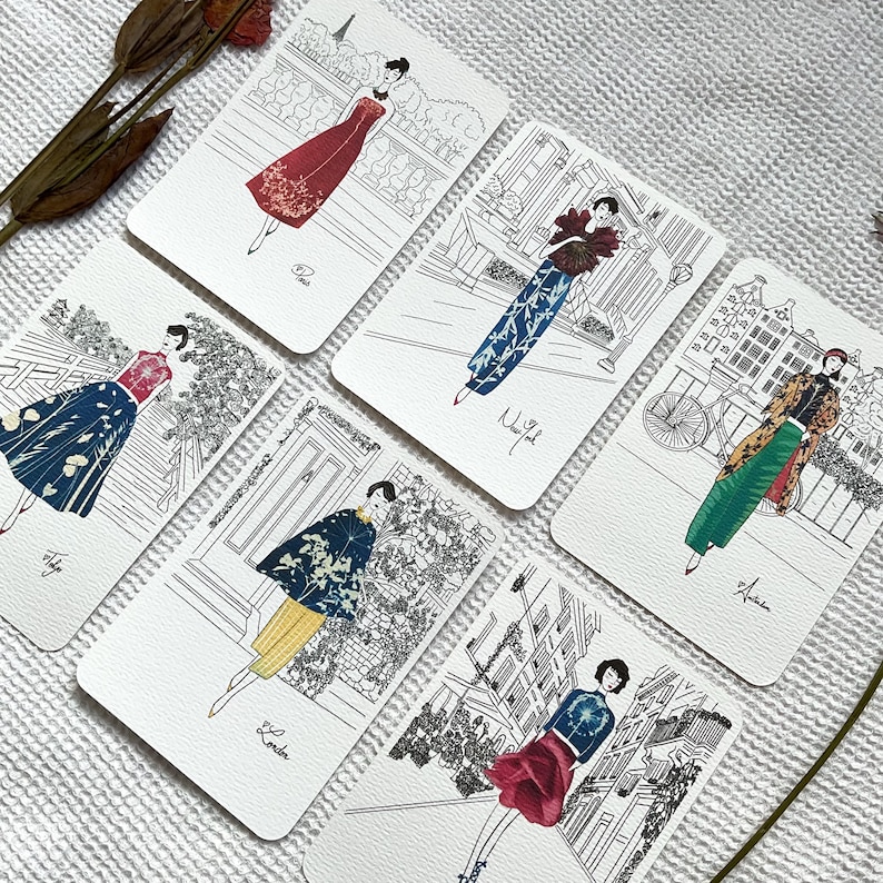 May include: Set of six illustrated cards featuring women in different outfits and locations. Each card has a black and white illustration of a woman in a different city, with the city name written below the illustration. The cities featured are Paris, New York, Amsterdam, London, Tokyo, and Rome.