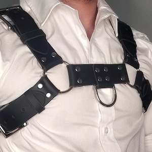 May include: Black leather chest harness with silver-tone metal hardware, including buckles, rings, and rivets. The harness features adjustable straps and a D-ring detail. The design is worn over a white collared shirt.
