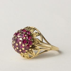 Op de afbeelding: Een gouden ring met een cluster van kleine, ronde, dieprode edelstenen. De edelstenen zijn gezet in een koepelvorm, omgeven door een gouden bloemenontwerp. De ring heeft een eenvoudige, gepolijste gouden band.