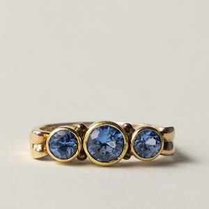 Könnte beinhalten: Ein goldener Ring mit drei runden, facettierten blauen Steinen in einer horizontalen Reihe. Der mittlere Stein ist größer als die beiden seitlichen Steine, und jeder ist in einer glatten, polierten Goldfassung auf einem massiven Goldring befestigt.