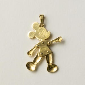 Puede incluir: Colgante dorado con la forma de Mickey Mouse. El colgante muestra a Mickey en una pose caminando con los brazos extendidos. La palabra "Disney" est&aacute; grabada en la parte posterior. Tiene un lazo en la parte superior para una cadena.