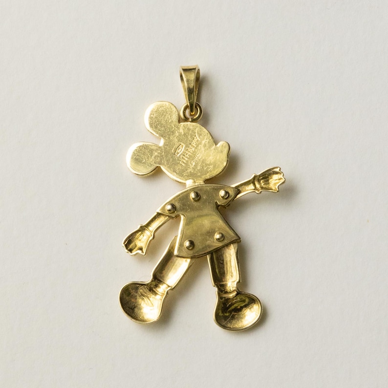 Puede incluir: Colgante dorado con la forma de Mickey Mouse. El colgante muestra a Mickey en una pose caminando con los brazos extendidos. La palabra "Disney" est&aacute; grabada en la parte posterior. Tiene un lazo en la parte superior para una cadena.