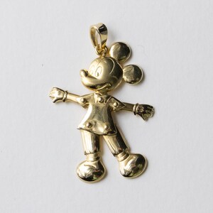 Puede incluir: Colgante dorado con forma de Mickey Mouse, con los brazos extendidos y un lazo para una cadena. El colgante es un dise&ntilde;o tridimensional detallado, con rasgos faciales y ropa visibles.