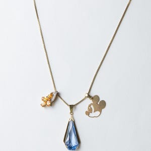 K&ouml;nnte beinhalten: Drei goldene Halsketten mit verschiedenen Anh&auml;ngern. Eine hat orangefarbene Edelsteine, eine andere einen blauen Kristall in Tropfenform und die dritte eine Mickey-Mouse-Silhouette. Auf wei&szlig;em Hintergrund.