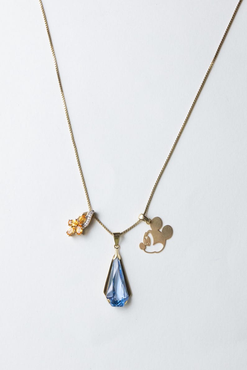 K&ouml;nnte beinhalten: Drei goldene Halsketten mit verschiedenen Anh&auml;ngern. Eine hat orangefarbene Edelsteine, eine andere einen blauen Kristall in Tropfenform und die dritte eine Mickey-Mouse-Silhouette. Auf wei&szlig;em Hintergrund.