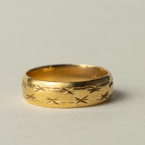 Könnte beinhalten: Ein goldener Bandring mit einem wiederkehrenden eingravierten Muster aus kleinen, sechsstrahligen Sternmotiven, die die Außenfläche umschließen. Das Metall hat eine polierte Oberfläche und der Ring hat eine gleichmäßige Breite.