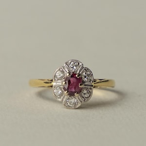 Op de afbeelding: Een gouden ring met een centrale ovaal geslepen dieprode robijn, omringd door zes kleine rond geslepen witte diamanten in een zilverkleurige bloemvormige zetting. De ringband is van glad goud met decoratieve uitgesneden details op de schouders.