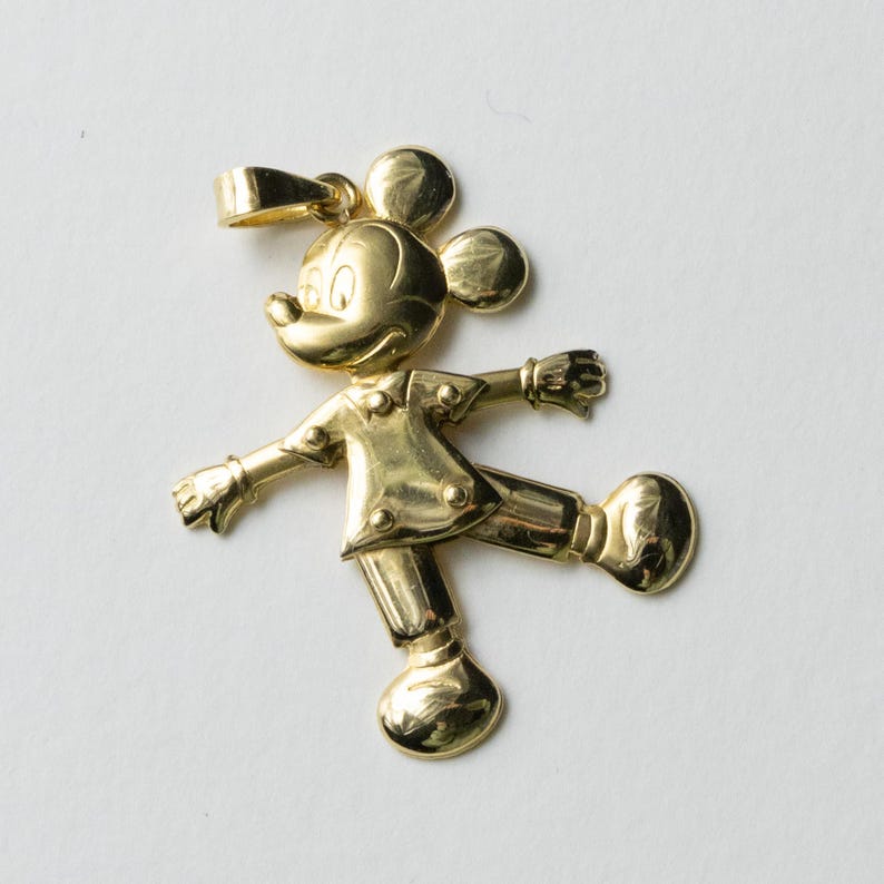 Puede incluir: Colgante dorado con la forma de Mickey Mouse, con los brazos y una pierna extendidos. El colgante tiene un lazo en la parte superior para una cadena. El personaje de dibujos animados lleva una camisa abotonada y pantalones.