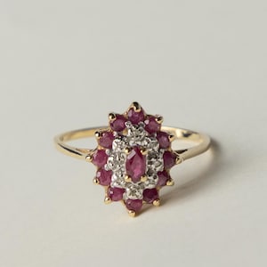 May include: A gold ring featuring a central marquise-cut ruby surrounded by a cluster of smaller round-cut rubies and tiny clear accent stones. The stones are set in a floral-shaped arrangement on a thin, polished gold band with a classic design.