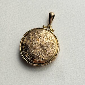 Puede incluir: Un colgante de medallón redondo en tono dorado con un intrincado patrón de volutas florales grabado en la superficie frontal. La carcasa de metal incluye una pequeña anilla en la parte superior. El texto 9CT BACKS & FRONTS R96239118 está grabado.