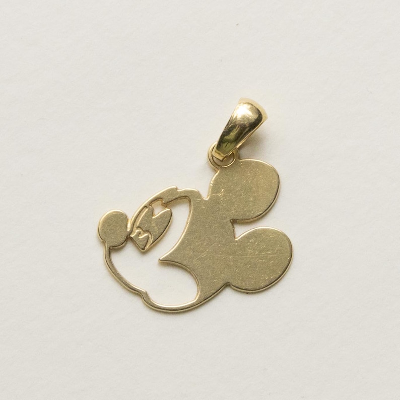K&ouml;nnte beinhalten: Ein goldfarbener Anh&auml;nger in Form von Mickey Mouse. Der Anh&auml;nger zeigt den Kopf und die Ohren der ikonischen Maus mit einer Schlaufe oben f&uuml;r eine Kette. Das Design ist schlicht und elegant, geeignet f&uuml;r eine Halskette oder ein Armband.