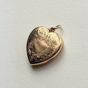 May include: A gold heart-shaped locket pendant featuring an intricate engraved floral pattern across the front surface. The locket includes a small circular jump ring at the top for attachment to a chain, showcasing a polished metallic finish.