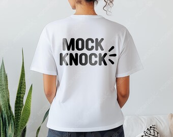 May include: White t-shirt with the text "MOCK KNOCK" in black, with a stylized sound effect graphic. The shirt is short-sleeved and appears to be made of a soft material. The model is wearing dark jeans.