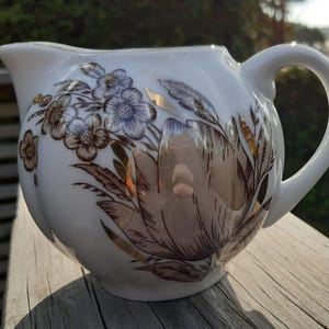 Może przedstawiać: Biała ceramiczna śmietanka z zaokrąglonym korpusem i zakrzywionym uchwytem. Śmietanka jest ozdobiona kwiatowym wzorem w odcieniach brązu, złota i niebieskiego. Śmietanka stoi na drewnianej powierzchni.